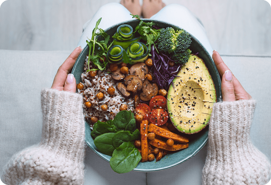 Person holding a bowl with avocado, broccoli, tomatoes, cucumber, purple cabbage, carrots, chickpeas, spinach, rice, and mushrooms on a light background.