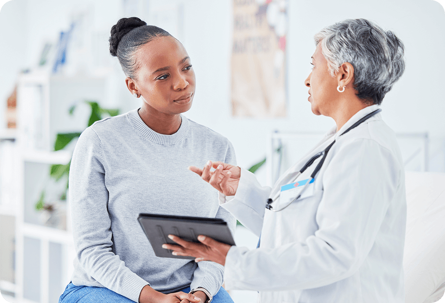 A doctor in a white coat holds a tablet and talks to a patient in a gray sweater seated on an examination table.