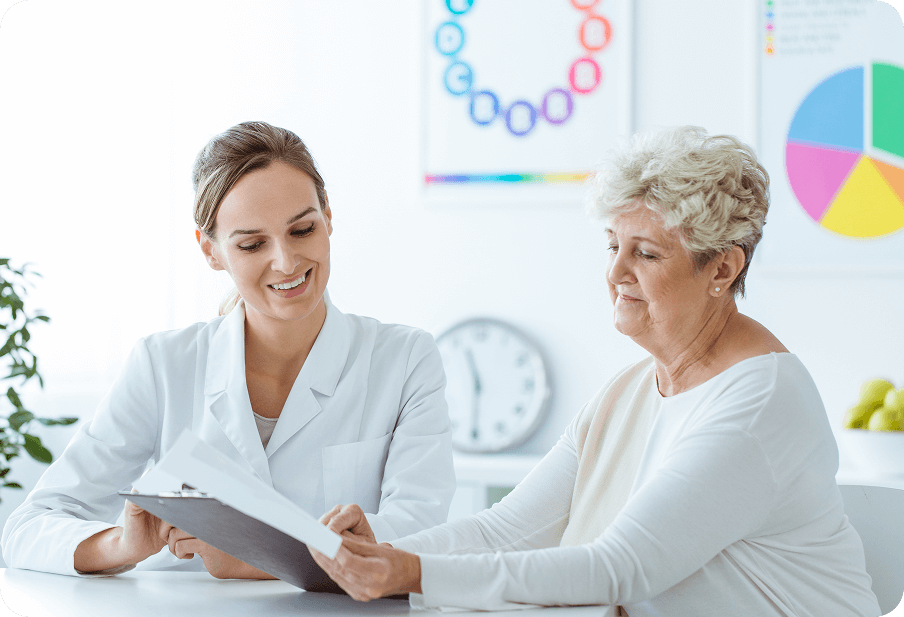 A medical professional discusses paperwork with an older woman in an office setting, featuring charts and a clock on the wall.